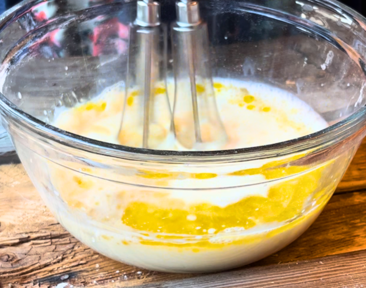 A woman beating a bowl with milk, oil, egg yolks in a glass bowl.