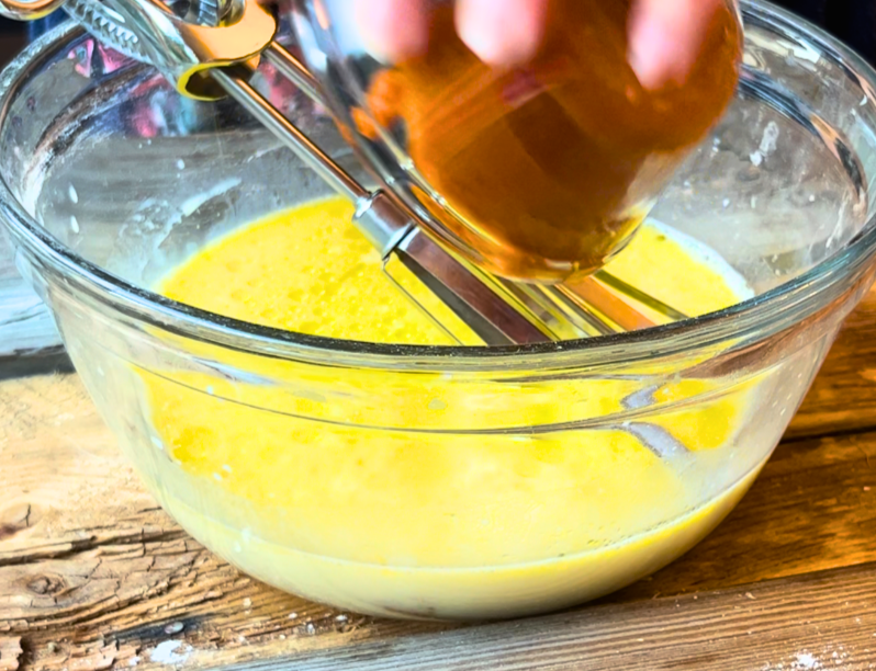 A woman adding pumpkin puree to a glass bowl. There are beaters in the bowl and yellow liquid.