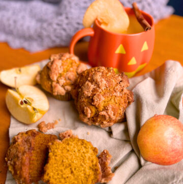 Pumpkin streusel muffins on a light green napkin with cut apples. One muffin in front is cut in half. The two whole muffins in the back are in front of a pumpkin shaped mug with hot apple cider. There is an apple slice on the rim on the mug and a cinnamon stick resting on top of the mug.