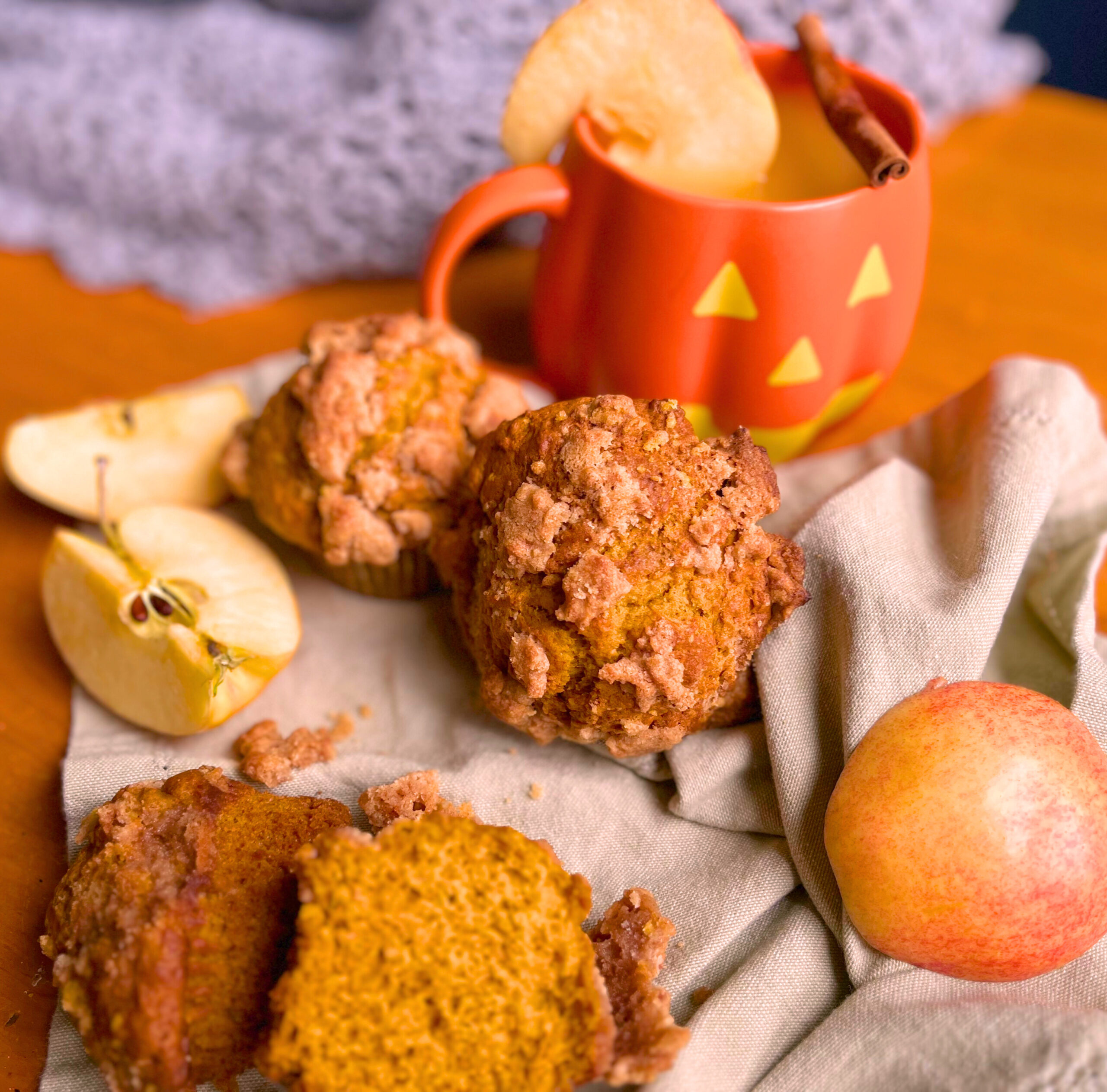 Pumpkin streusel muffins on a light green napkin with cut apples. One muffin in front is cut in half. The two whole muffins in the back are in front of a pumpkin shaped mug with hot apple cider. There is an apple slice on the rim on the mug and a cinnamon stick resting on top of the mug.
