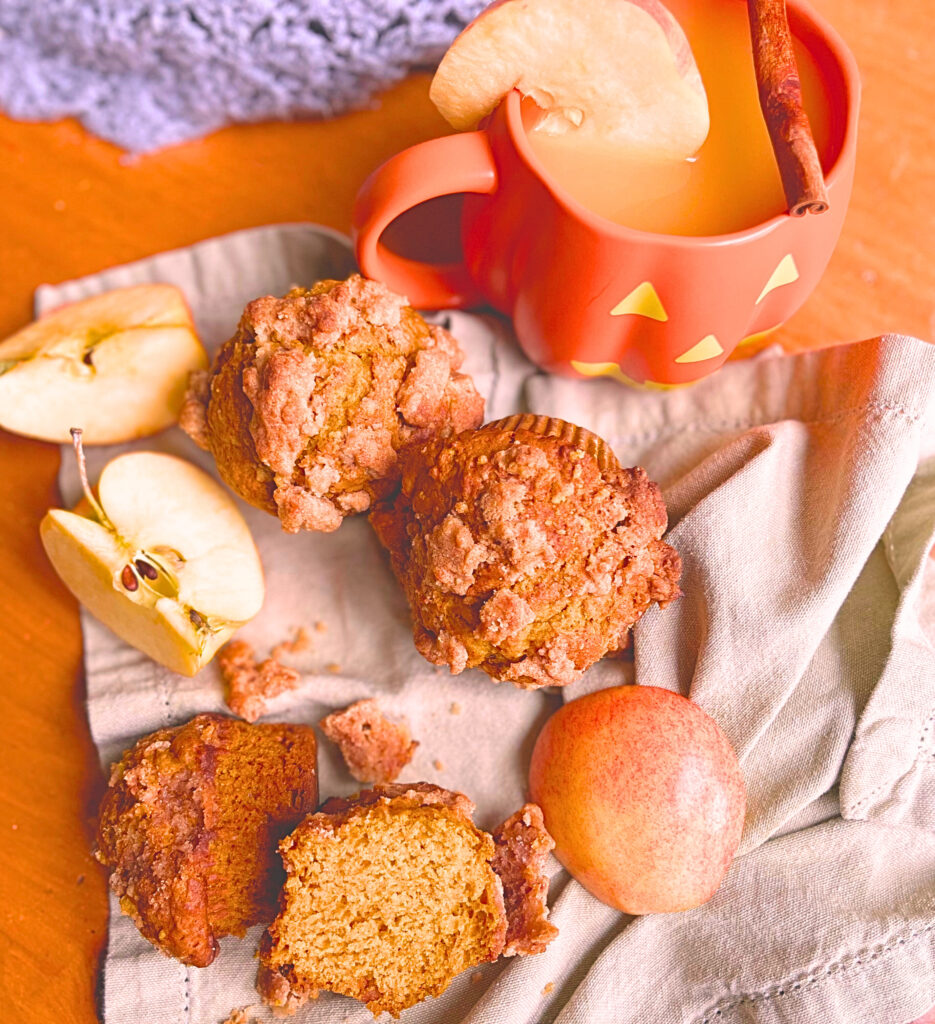 Pumpkin streusel muffins on a light green napkin with cut apples. A pumpkin shaped mug with hot cider, an apple slice on the rim, and a cinnamon stick resting on top.