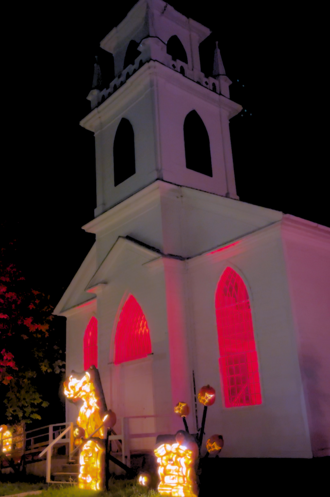A white church at night, with red glowing lights from inside. Decorated and lite up pumpkins in front.