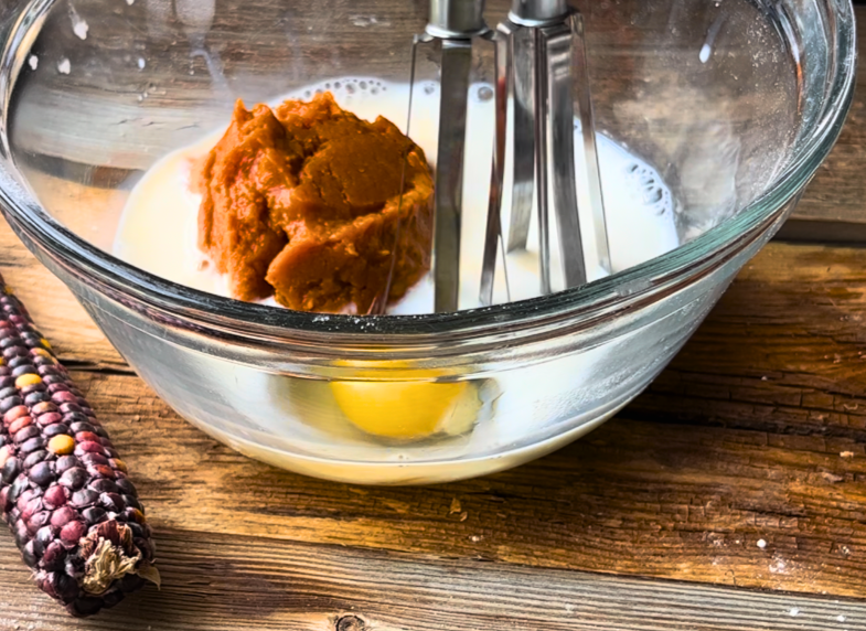 A glass bowl with milk, egg, and cooked pumpkin. A rotary beater in the bowl.