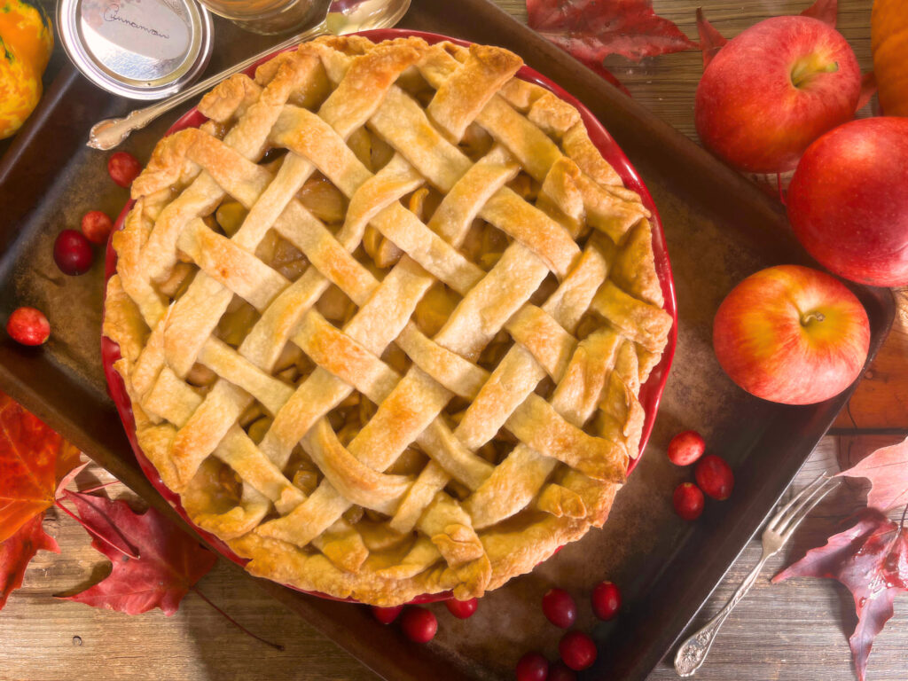 An apple pie with a lattice work top crust. Three apples sitting beside and a jar of cinnamon on the opposite side. A silver spoon and fork beside the pie. All on a brown tray surrounded by cranberries.