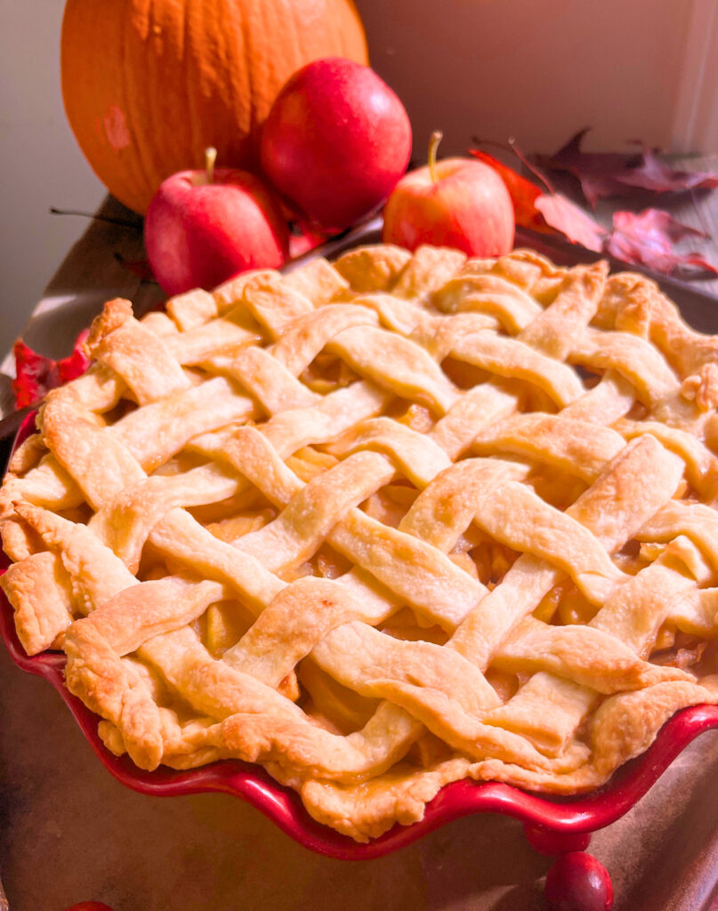 A baked apple pie with a lattice work top, in a red pie plate. A pumpkin and apples in the background