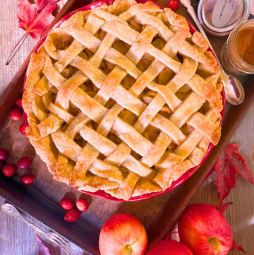 An apple pie with a lattice work top with apple in the foreground a spoon and a jar of cinnamon in the back ground