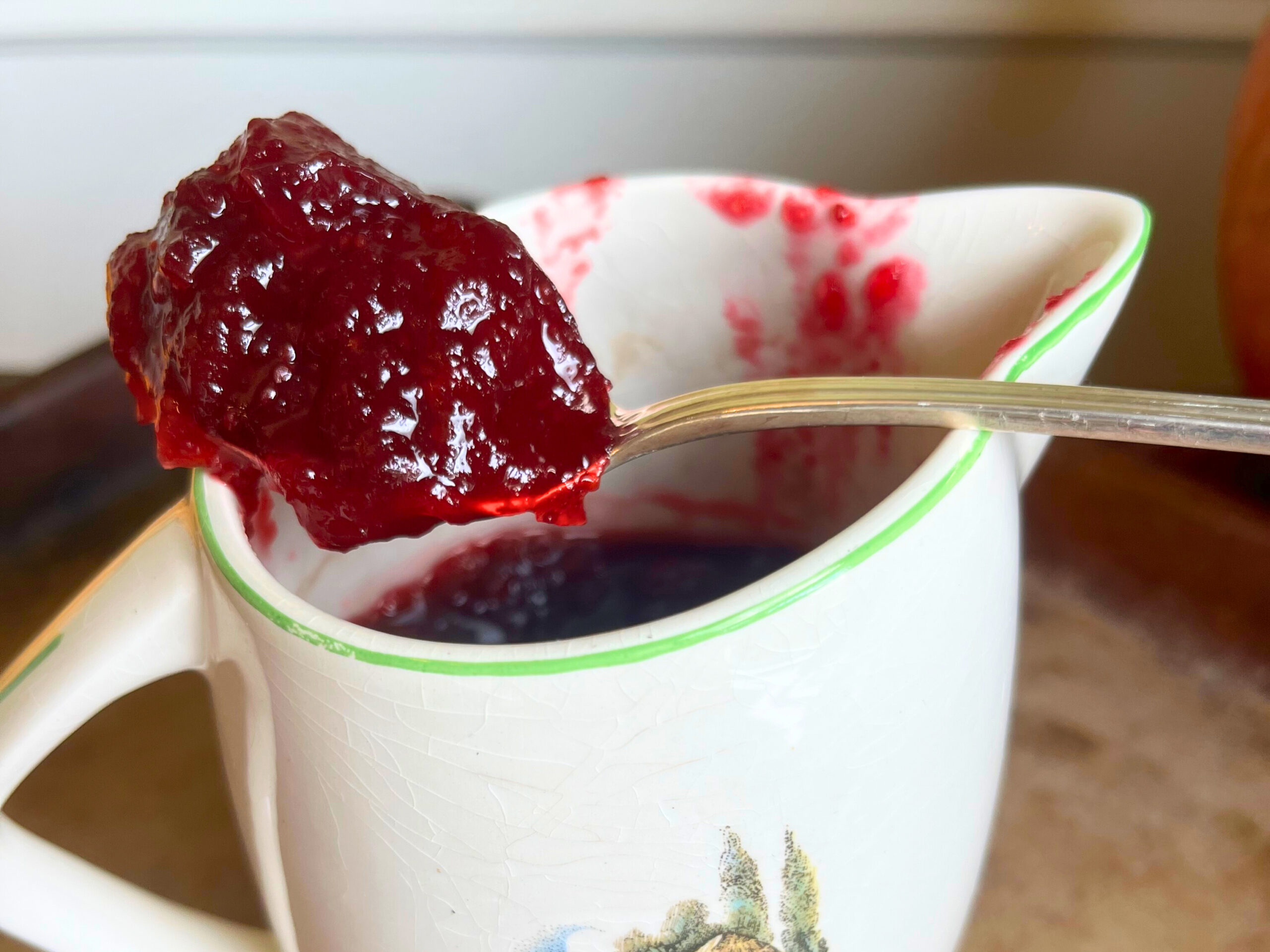 A small with and green pitcher with cranberry sauce in it. A spoon resting on tip with a scoop of cranberry sauce.