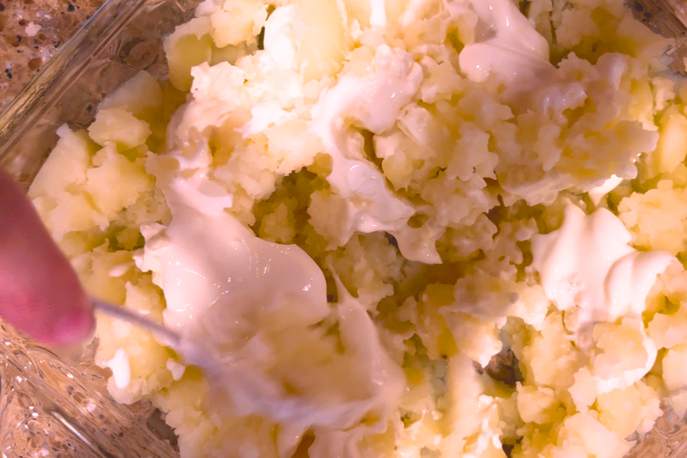 Mashed potatoes in a square, glass casserole dish with cream cheese, sour cream and butter being mixed in.