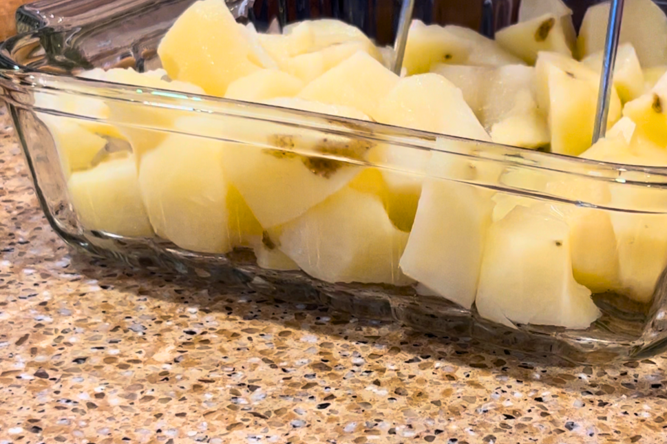 A square, glass casserole dish with peeled, cut, and cooked potatoes being mashed with a potato masher