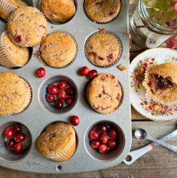 A muffin tin with cranberry sauce muffins. Some fresh cranberries on the tray and a plate with a partly eaten muffin. Two measuring spoons in front and a jar of hydrangeas in the background