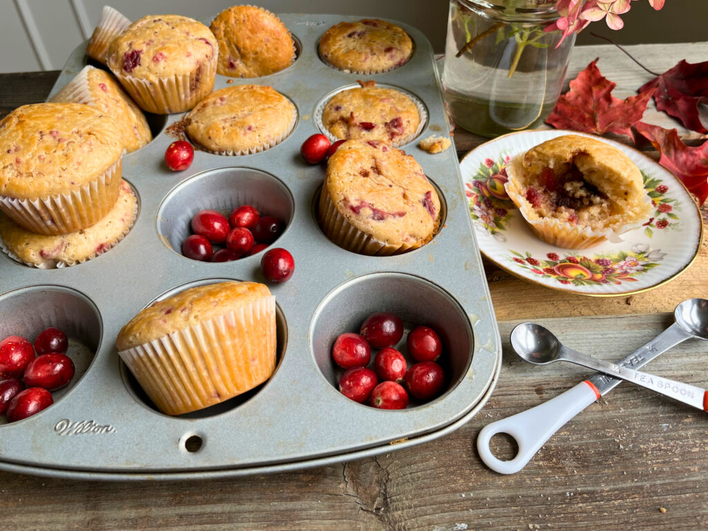 Cooked cranberry muffins in a muffin tray. Some fresh cranberries around them and a plate with a partly eaten cranberry muffin. Two measuring spoons in the foreground.