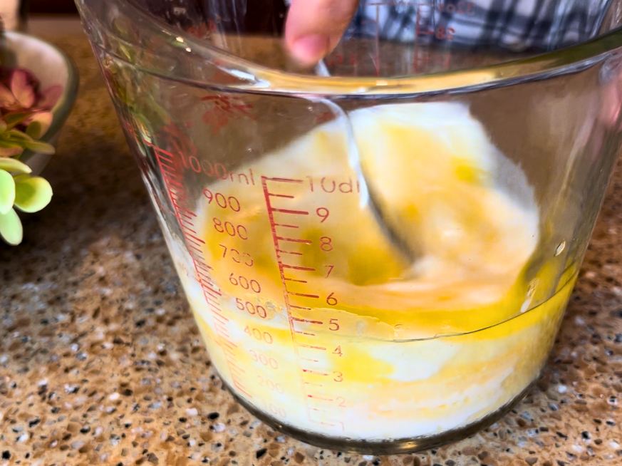 Woman mixing milk, egg, and melted butter in a large glass measuring cup, with a fork.