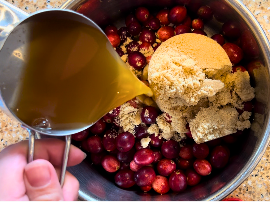 Woman pouring apple cider from a metal measuring cup into a sauce pot with cranberries and brown sugar