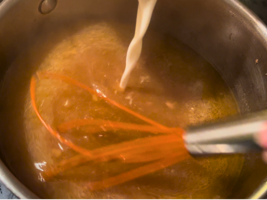 Pouring cornstarch mixture into a pot of boiling turkey drippings. An orange whisk inside the pot.