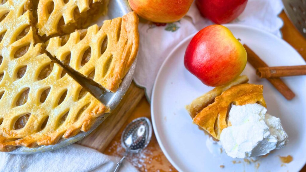 A sliced apple pie. There is one slice on a white plate with whipped cream on top.