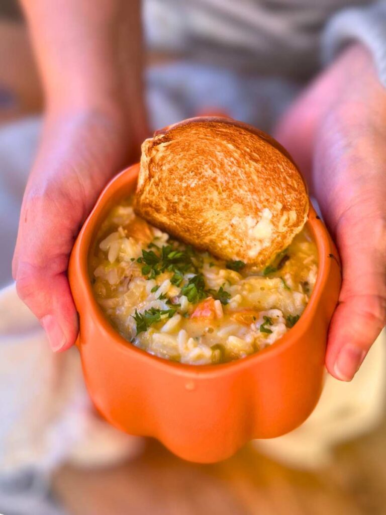 A woman is holding an orange pumpkin shaped mug filled with turkey and rice soup. There is a slice of toast inside.