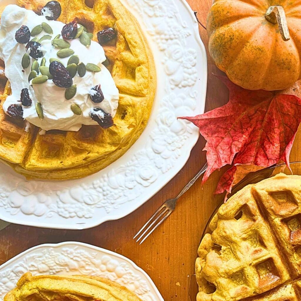 Three stacks of pumpkin waffles. One is topped with whipped cream, pumpkin seeds, and dried cranberries.