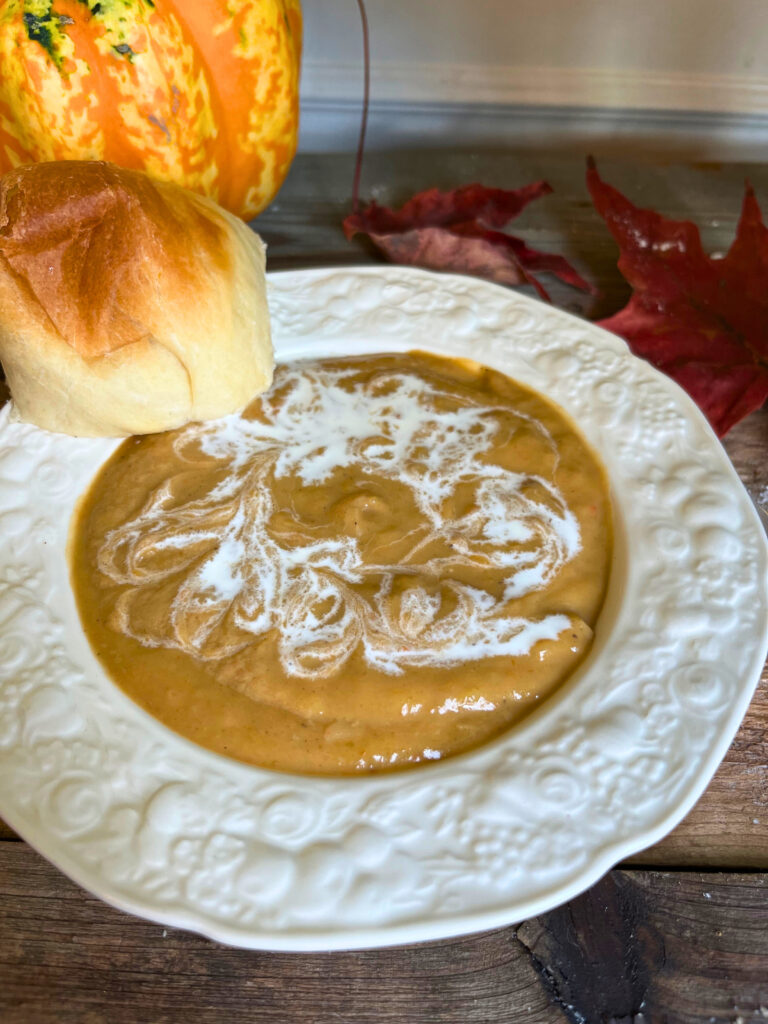 A white bowl of butternut squash soup , with a swirl of cream on top. A roll off to the side. A gourd and maple leaves in the background.