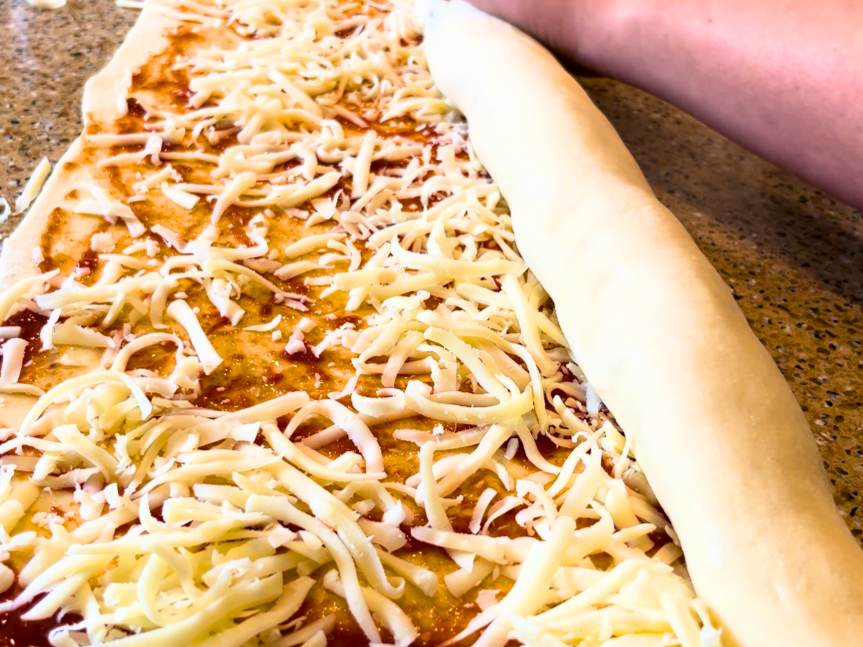 A woman rolling bread dough, with pizza sauce and cheese, into a log shape