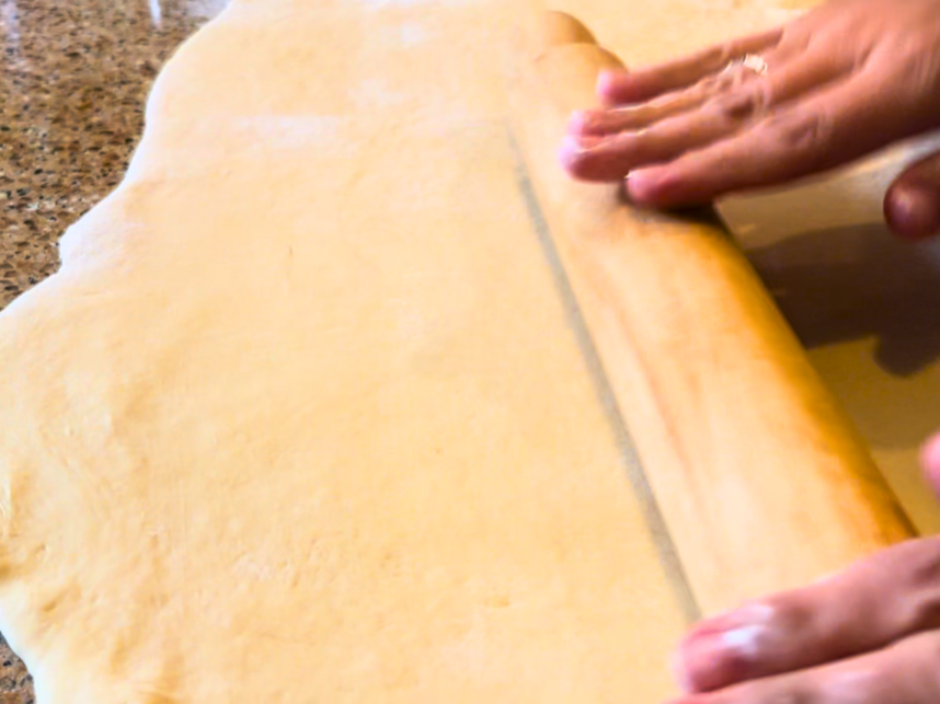A woman using a wooden rolling pin to roll bread dough into a rectangle