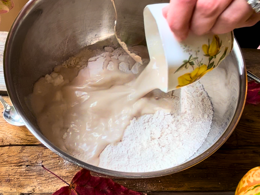 Pouring a yeast mixture into a metal bowl with flour in it.
