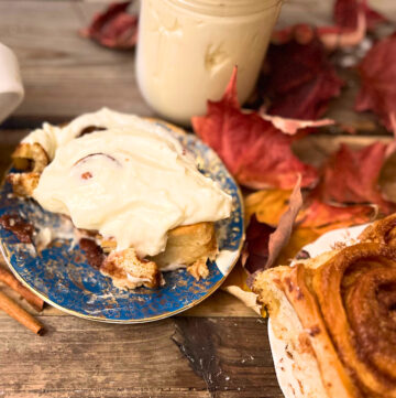A cinnamon roll with cream cheese icing on a blue and gold plate. An cinnamon roll without icing in the foreground. A jar off icing and a cup off coffee off to the side