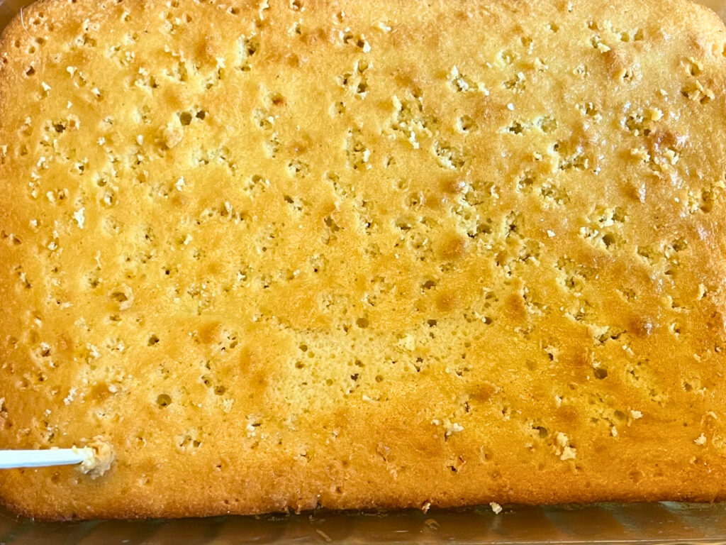 A overhead view of a sponge cake with a skewer punching holes in it.
