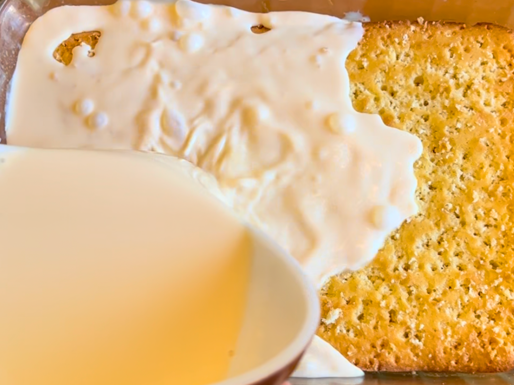 Pouring a milk mixture on top of a cooked sponge cake with holes poked in it, in a glass casserole dish.