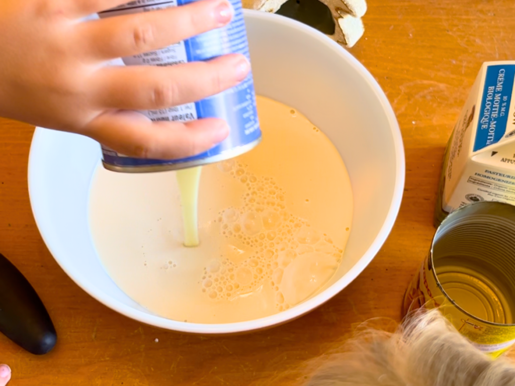 Child adding sweetened condensed milk to a porcelain bowl with cream and evaporated milk in it.