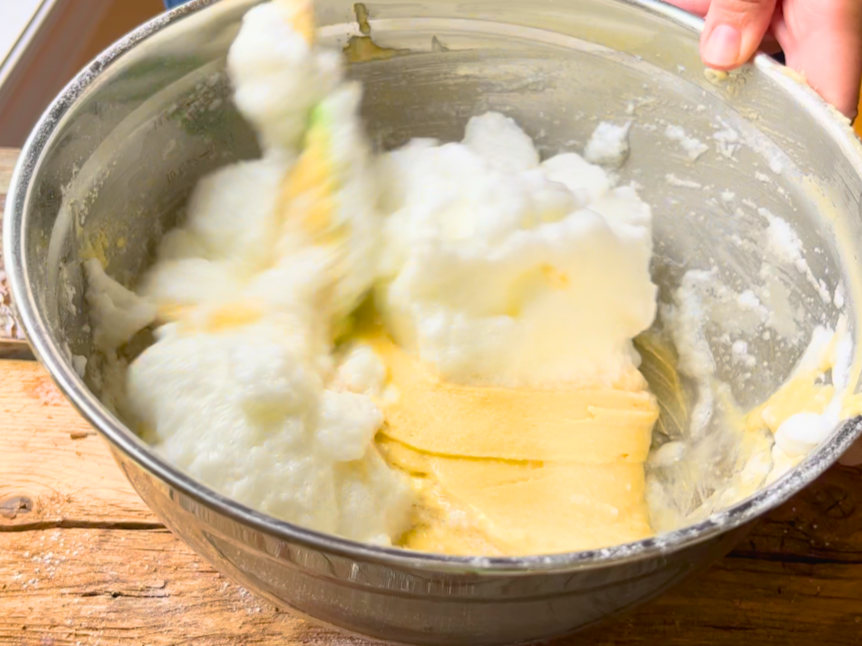 Woman folding the egg whites into the cake batter, with a spatula in a metal bowl.
