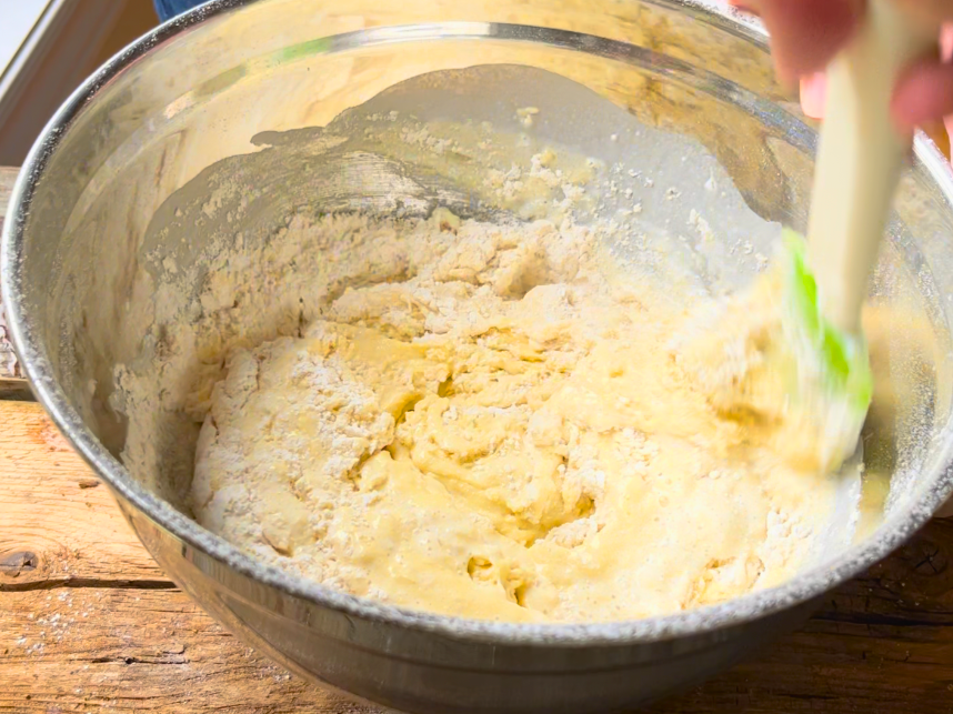 Mixing an egg yolk mixture with a sifted flour mixture, in a metal bowl with a green and white spatula.