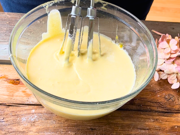 Woman beating an egg yolk, sugar, and vanilla mixture in a glass bowl with rotary beaters.