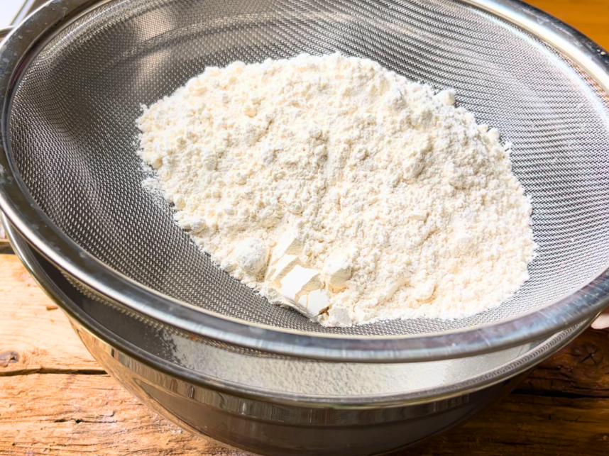Flour mixture in a sieve on top of a large metal bowl.