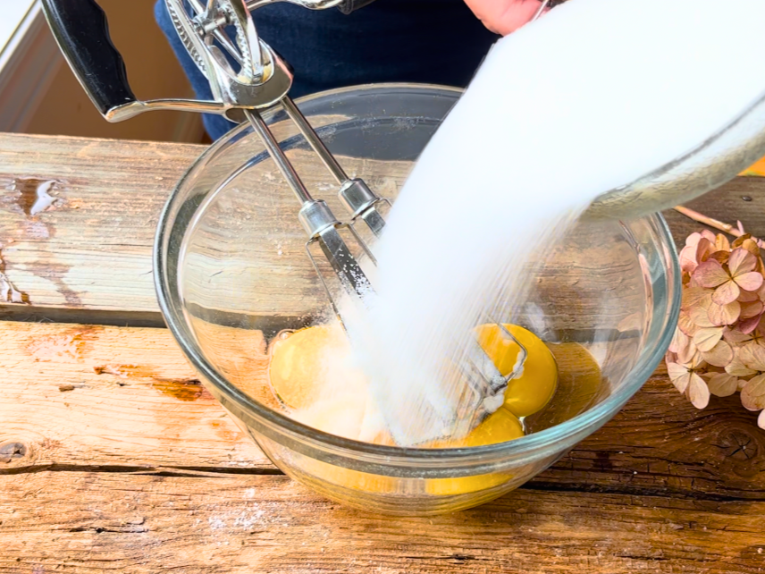 Woman pouring sugar on top of a glass bowl with egg yolks in it.