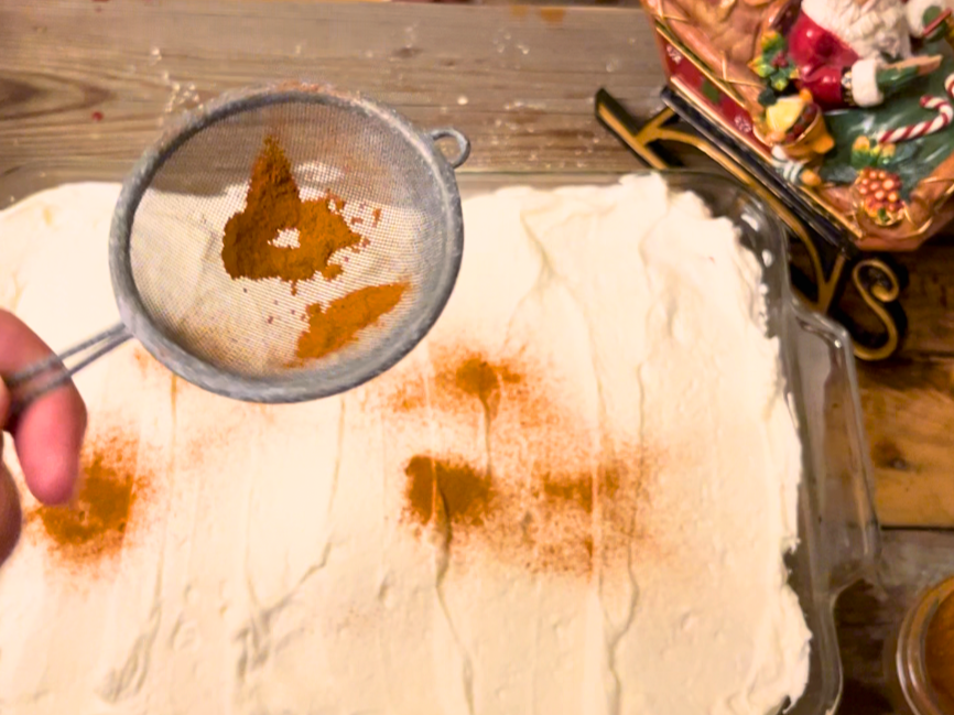 Woman dusting a tres leches cake with cinnamon using a small sifter.