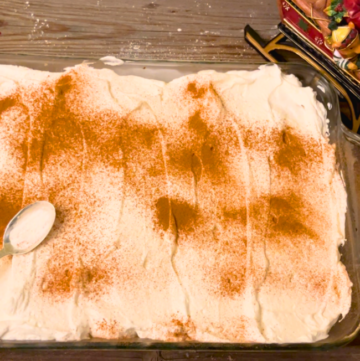 Tres leches cake on a wooden table with cinnamon sprinkled on top. A metal spoon resting on top of the cake.
