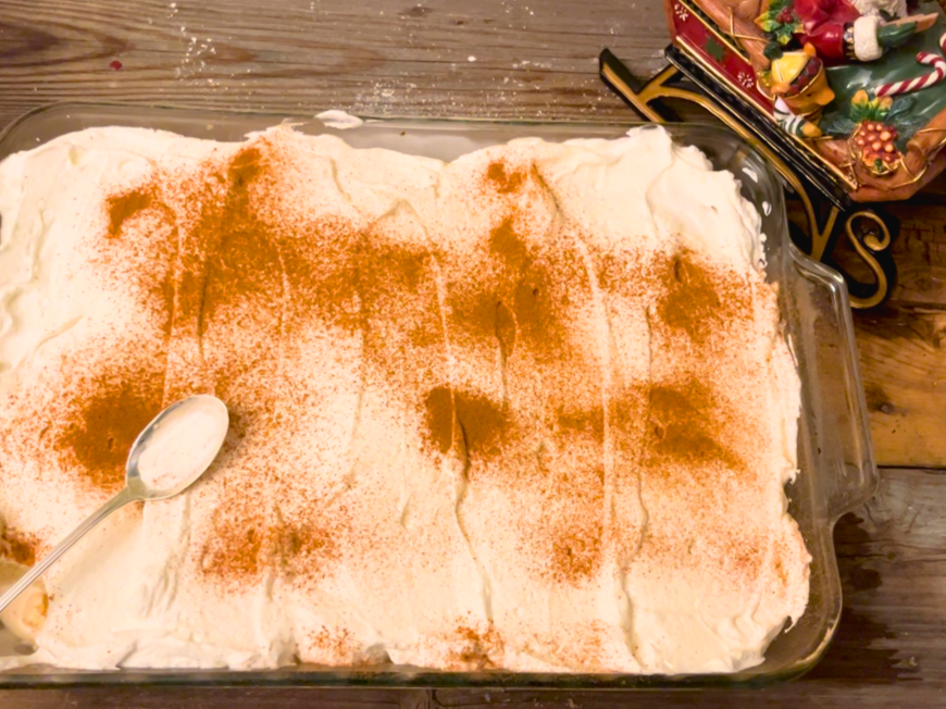Tres leches cake on a wooden table with cinnamon sprinkled on top. A metal spoon resting on top of the cake.
