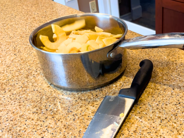 A metal sauce pot with peeled and sliced apples inside. A kitchen knife in the foreground