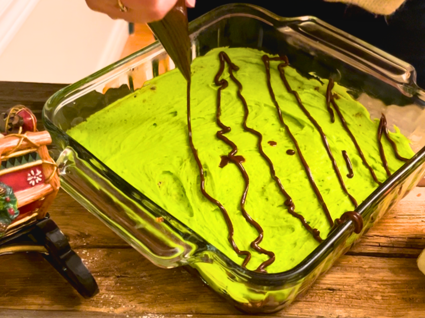 Woman adding a mint chocolate drizzle, using a pipping bag, on top of a square casserole dish of no-bake chocolate mint bars