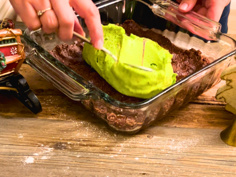 A woman spreading green mint icing onto a chocolate base in side of a square casserole dish.