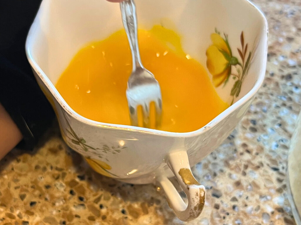 A beaten egg in a white and yellow flowered cup, with a metal fork.