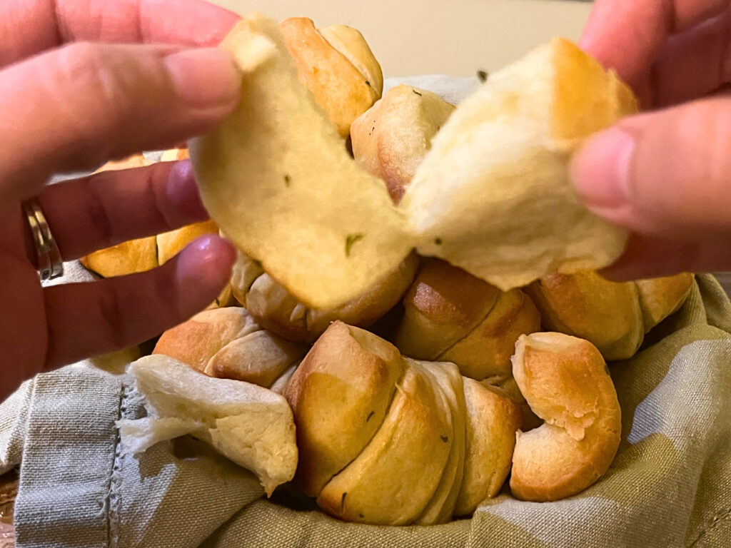 A woman holding and pulling apart a garlic pull apart dinner roll. A basket of dinner rolls in the background.