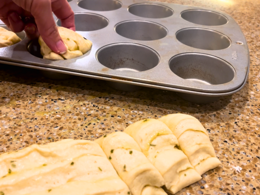 A long strip of dough cut into portions and being placed into a muffin tin.