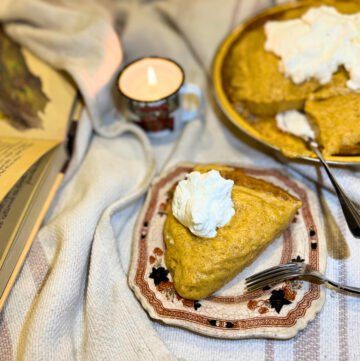 A piece of pumpkin chiffon pie in the foreground. The full pie in the background beside a lite candle. An open book off to the side. All on a cream blanket.