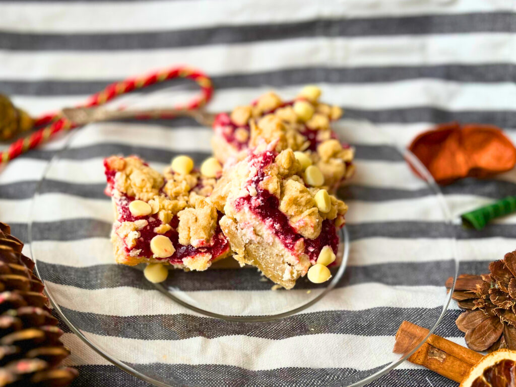 Three cranberry cheesecake bars on a glass plate sitting on a grey and white cloth