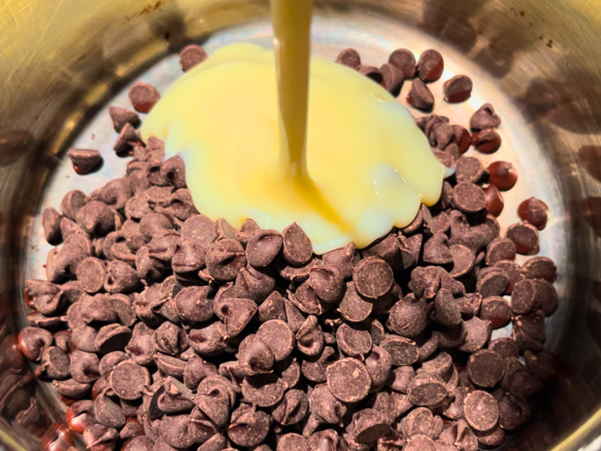 Sweetened condensed milk being poured over a pot with chocolate chips in side.