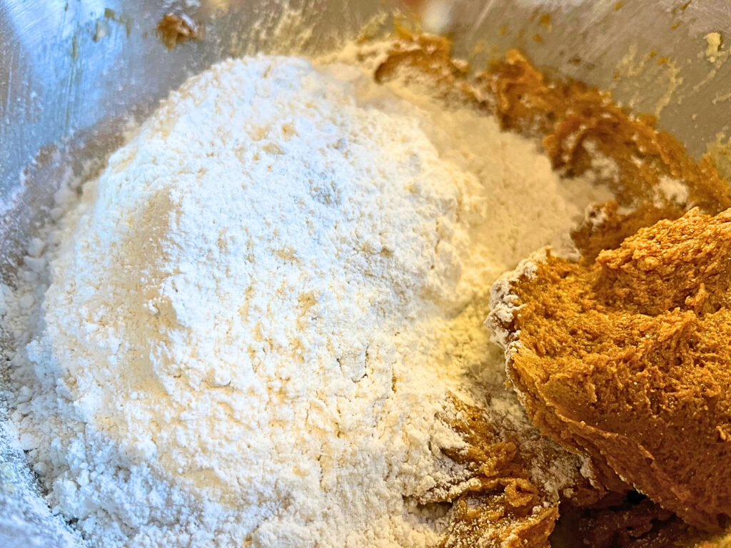 A gingerbread cookie mixture in a metal bowl with flour being added.