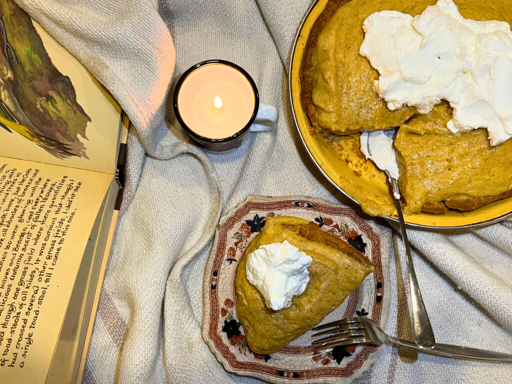 A piece of pumpkin chiffon pie in the foreground with the full pie behind it. A candle burning in the middle of the frame. An open book off to the side. All on a cream blanket.