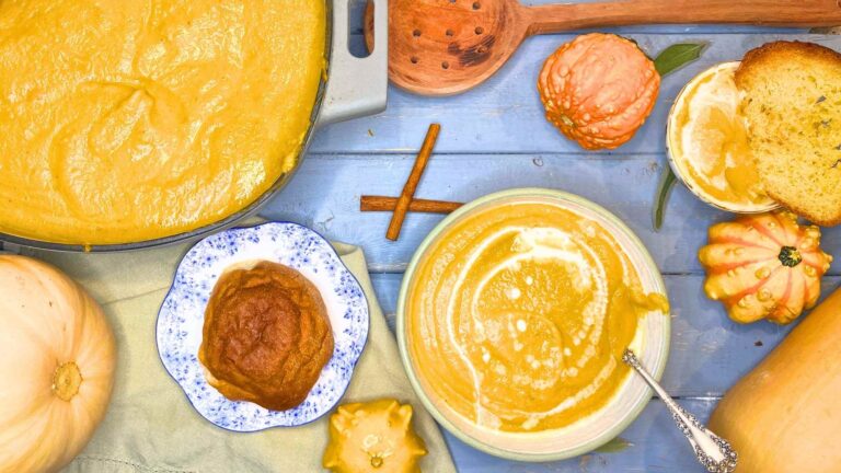 A bowl of butternut squash soup. There is a smaller one off to the side with a slice of toast dipped in it. There is a dinner roll and a large pot of soup.