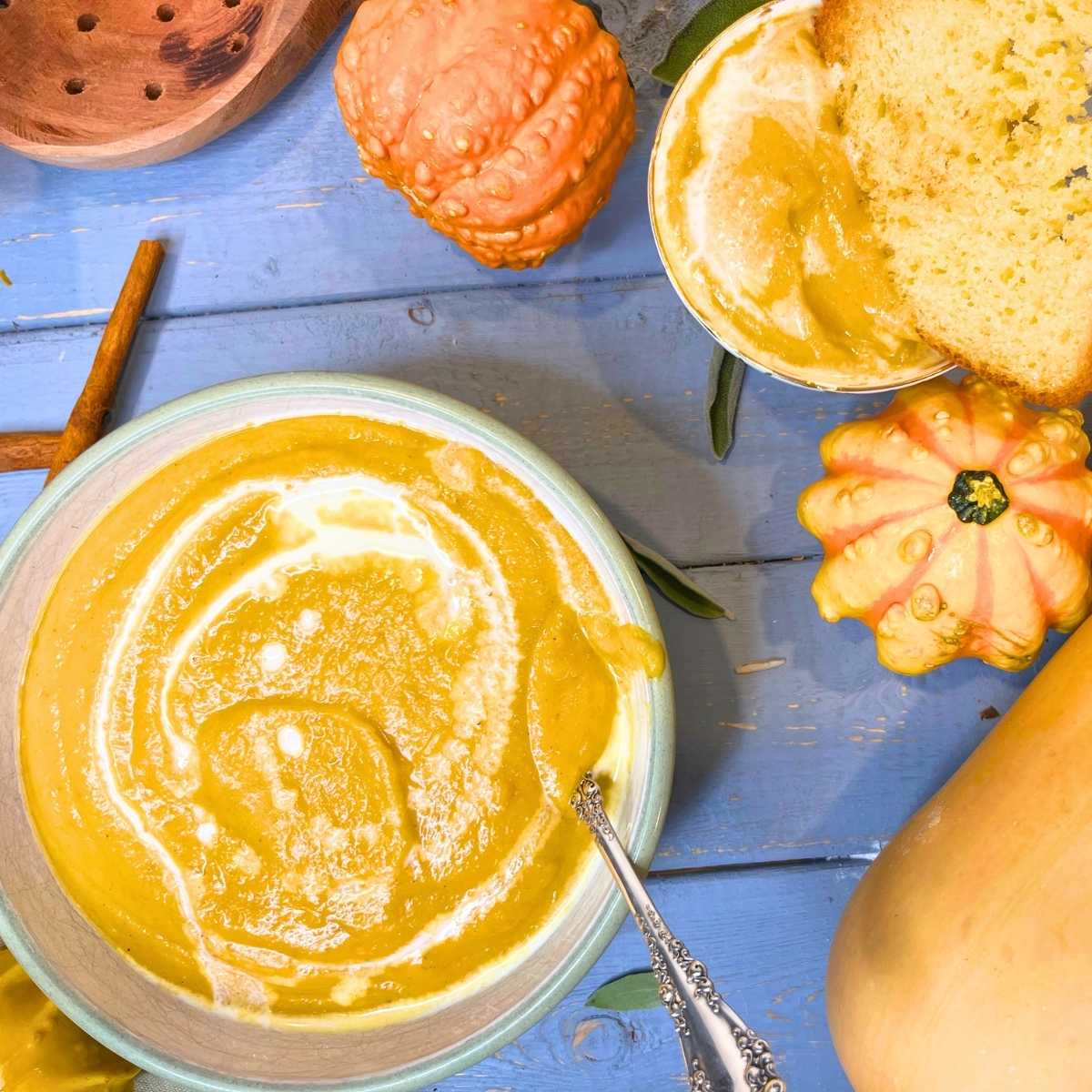 A bowl of butternut squash soup with a spoon inside and a swirl of cream on top. There is a small bowl of soup off to the side with a slice of toast inside.